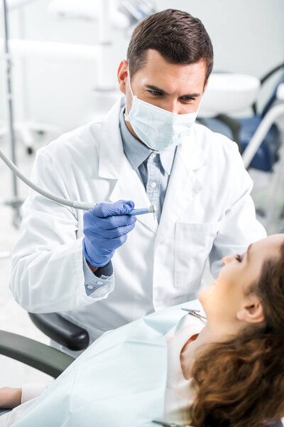 селективный фокус стоматолога в белом халате и маске, держащего стоматологический инструмент рядом с пациенткой
