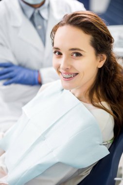 selective focus of woman in braces smiling with dentist standing with crossed arms on background clipart