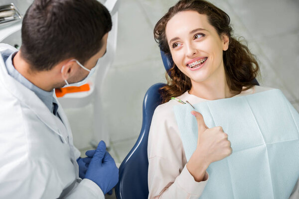 selective focus of woman in braces smiling while showing thumb up near dentist during examination 