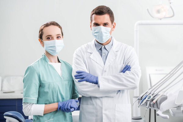 female dentist in mask standing near coworker with crossed arms in dental clinic