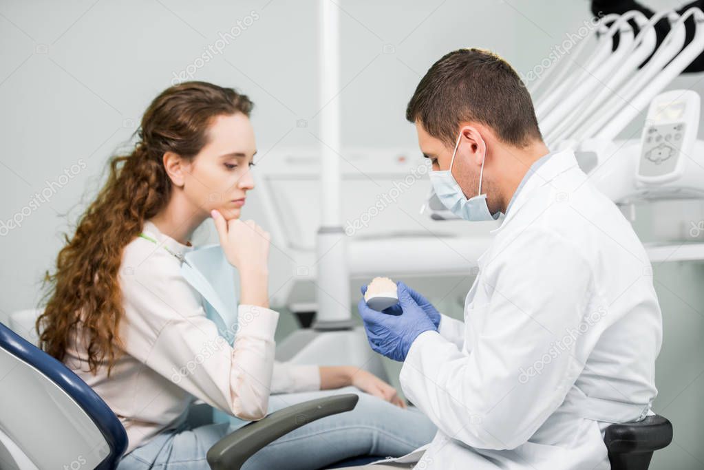 dentist in latex gloves and mask holding teeth model near pensive woman 