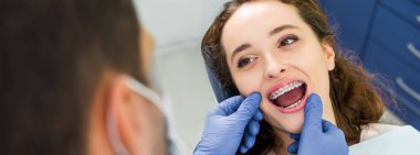 selective focus of cheerful woman in braces opening mouth during examination of teeth near dentist clipart