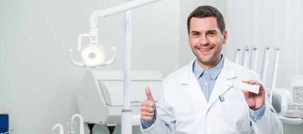 handsome dentist holding toothpaste and showing thumb up in dental clinic