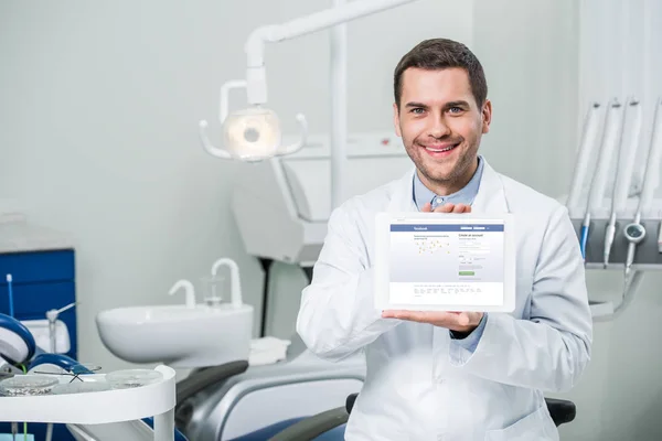 愉快的牙医拿着数字平板电脑与脸书应用程序在屏幕上的牙科诊所 — 图库照片