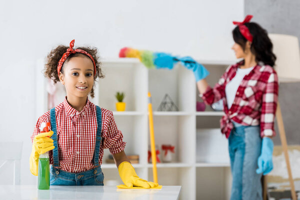 selective focus of smiling african american child cleaning table while mother dusting shelving unit