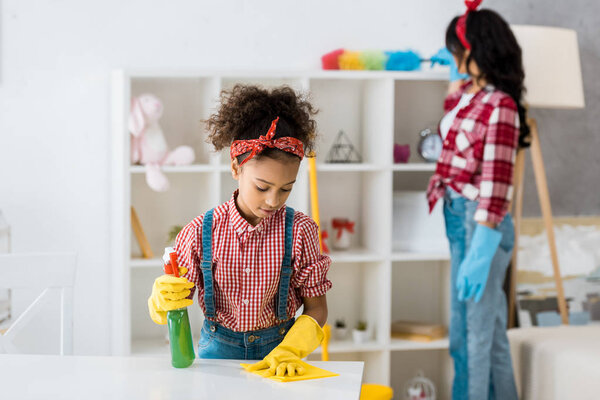 selective focus of cute african american child cleaning table while mother dusting furniture