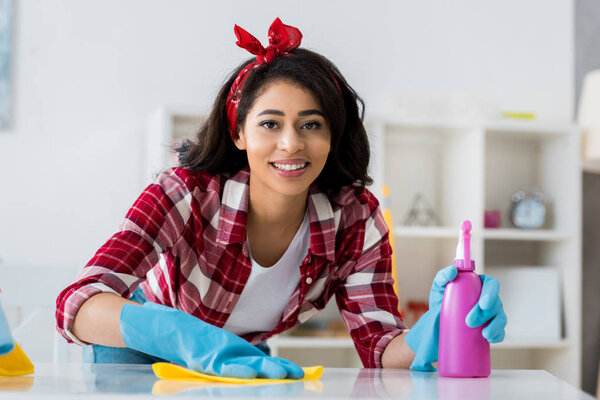 улыбающаяся африканская американка с желтой тряпкой и розовой бутылкой, смотрящей в камеру
