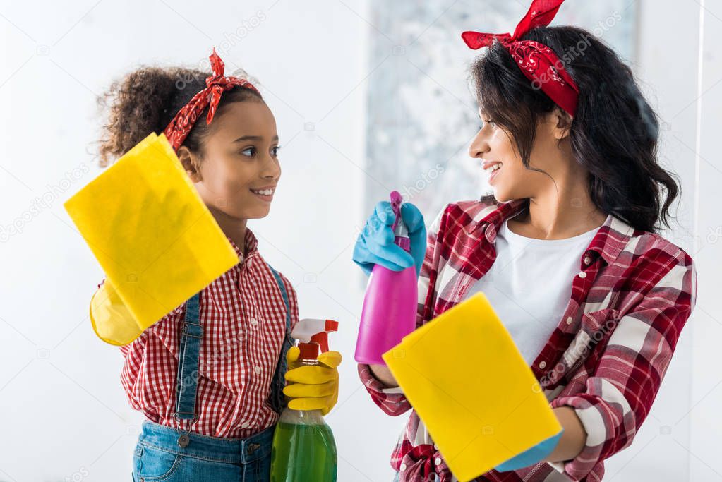 smiling african american mom and daughter with yellow rags and spray bottles cleaning house