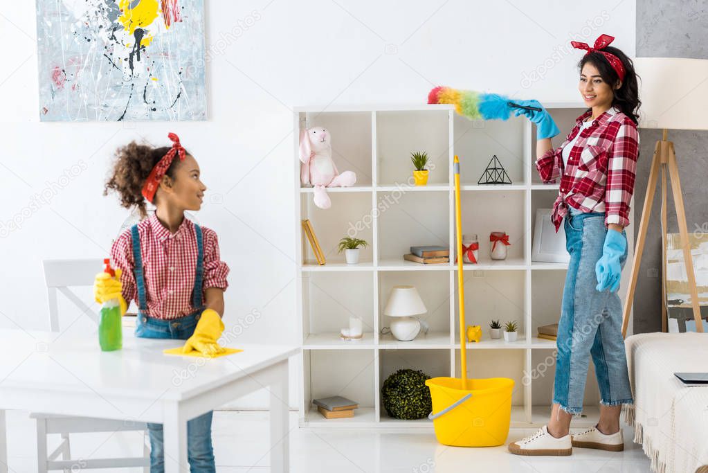 cute african american child cleaning table while mother dusting shelving unit