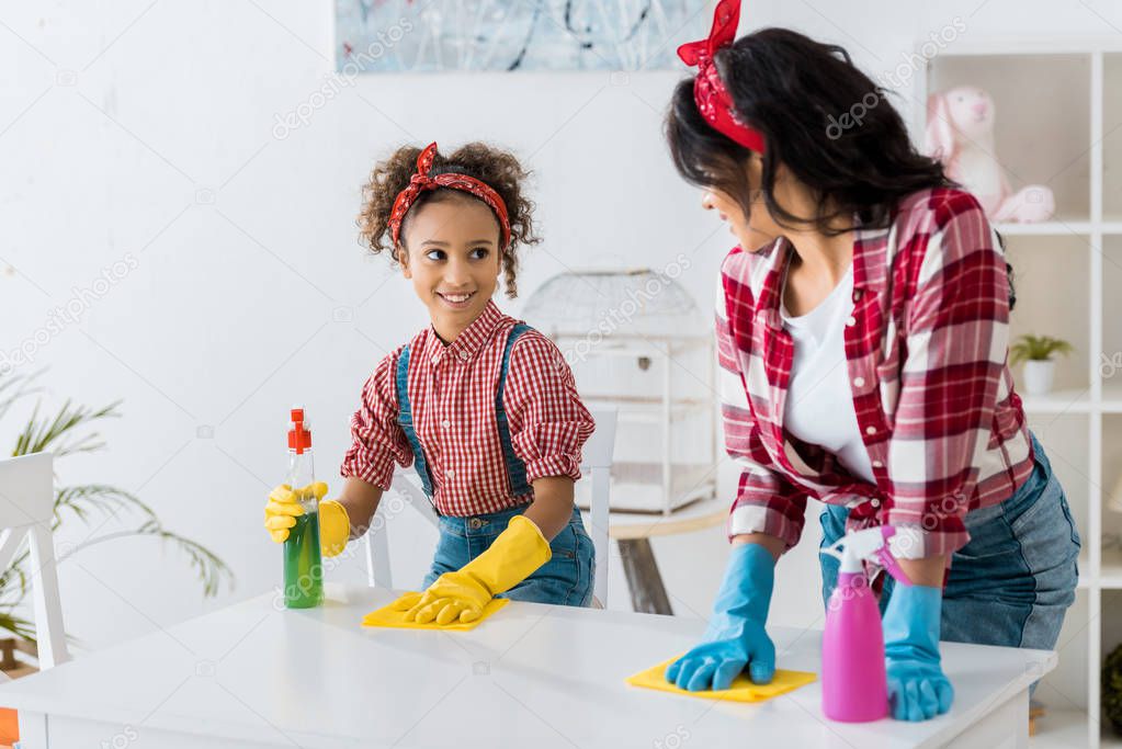 smiling african american mom and daughter cleaning white table with yellow rags