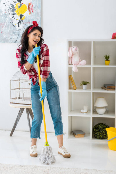 beautiful african american woman in plaid shirt and blue rubber gloves singing with mop