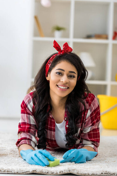 улыбающаяся африканская американка в синих резиновых перчатках чистит ковер губкой
