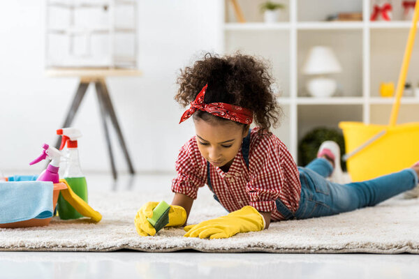 selective focus of cute african american girl lying on carpet and cleaning it with sponge