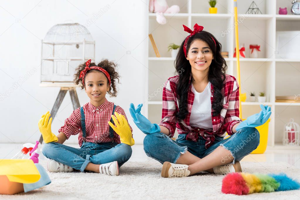 smiling african american mom and daughter and plaid shirts and bright rubber gloves sitting in lotus pose 