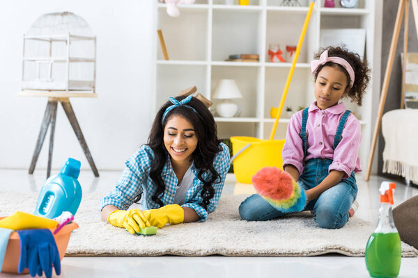 cute african american child sitting on carpet with dusting brush while mother cleaning carpet