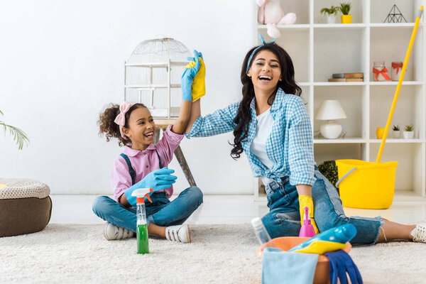 laughing african american mom holding daughters raised hand while sitting on carpet