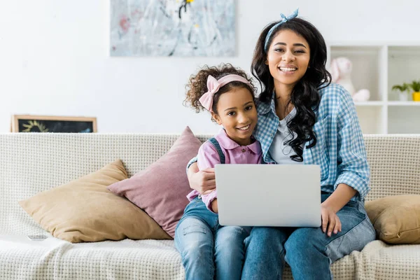 アフリカ系アメリカ人の母と娘がソファに座ってノート パソコンを使用して笑みを浮かべてください — ストック写真