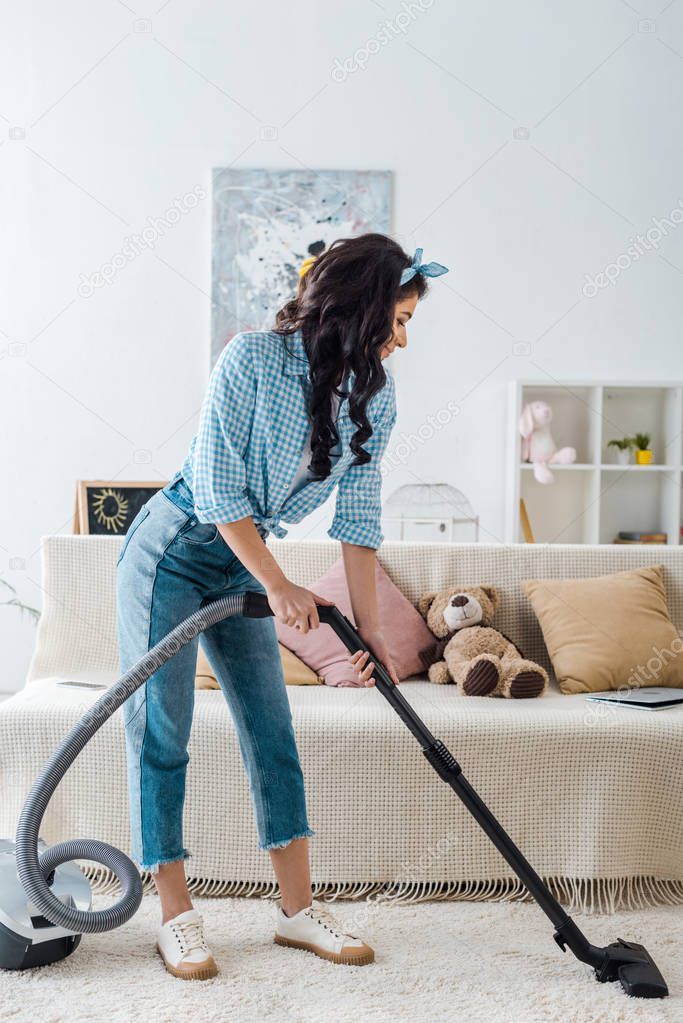 african american woman in plaid shirt and blue jeans vacuuming carpet