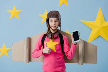 Neşeli çocuk smartphone boş ekran ile tutarak ve başparmak mavi yıldızlı arka plan üzerinde gösterilen uçuş kask içinde