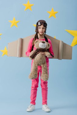 Oyuncak ayı mavi yıldızlı arka plan üzerinde tutarak karton kanatlı meraklı çocuk