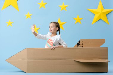 Kid with braids waving hand while sitting in cardboard rocket on blue starry background clipart
