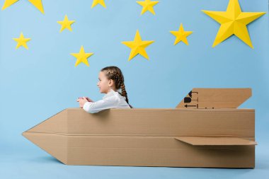 Side view of smiling kid with gamepad sitting near cardboard rocket on blue starry background clipart