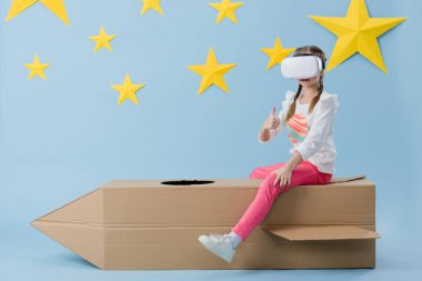 Karton roket üzerinde oturan ve mavi yıldızlı arka plan üzerinde başparmak gösterilmesini vr kulaklık evlat