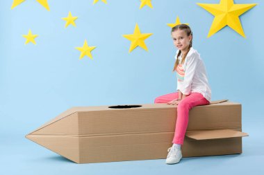 Mavi yıldızlı zemin üzerine karton roket oturup örgü ile gülümseyen çocuk