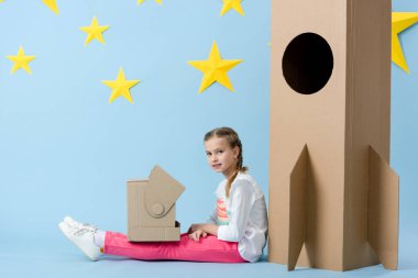 Karton roket oturan ve kameraya mavi yıldızlı arka plan üzerinde seyir kask ile baktılar çocuk