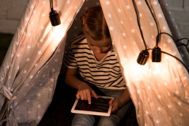 Child in striped t-shirt sitting in wigwam and using digital tablet with blank screen clipart