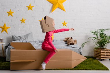 Funny kid in cardboard helmet sitting on rocket with hand up in bedroom clipart