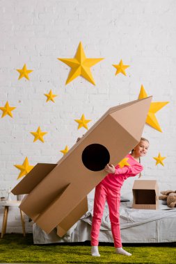 Full length view of excited kid holding big cardboard rocket in bedroom clipart
