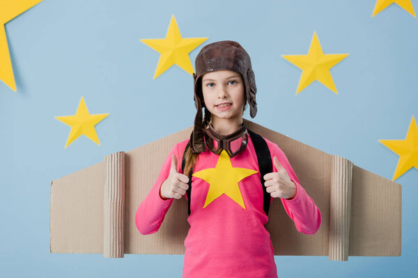 Kid in flight helmet and cardboard wings showing thumbs up on blue starry background