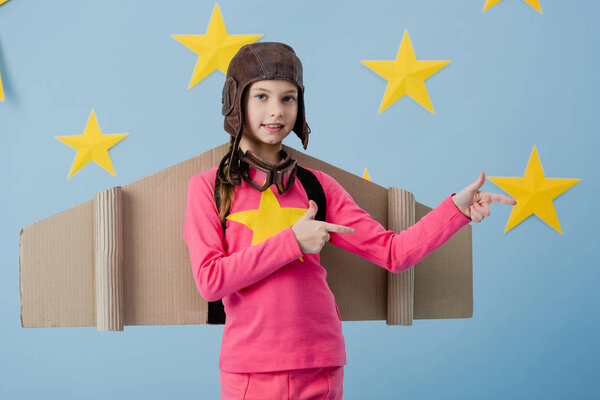 Smiling child with cardboard wings pointing with fingers on blue starry background