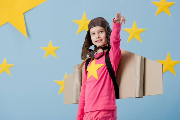 Excited kid in pilot costume pointing with fingers on blue background with stars