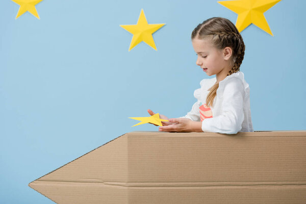 Child with braids sitting in cardboard rocket and holding yellow star on blue starry background