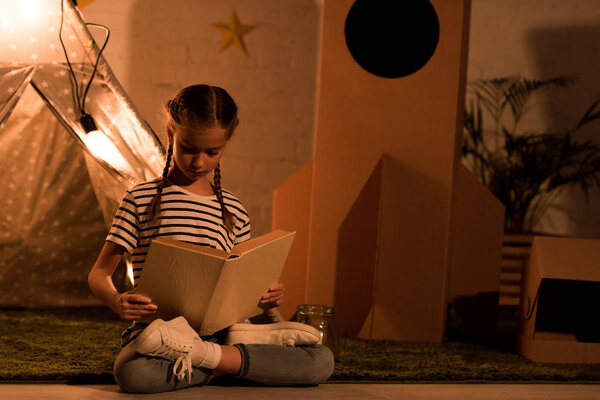 Ребенок-подросток, сидящий в позе лотоса и читающий книгу в темной комнате
