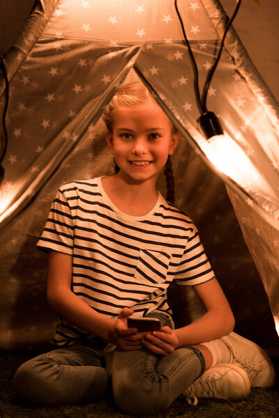 Laughing kid in striped t-shirt sitting in wigwam and holding smartphone