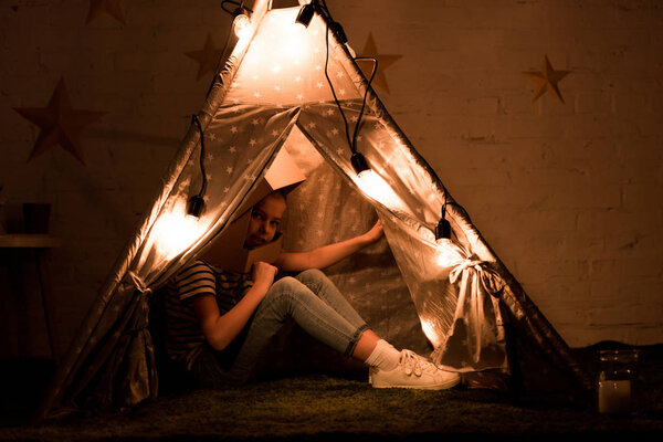 Preteen kid sitting in cozy wigwam with bulbs in dark room