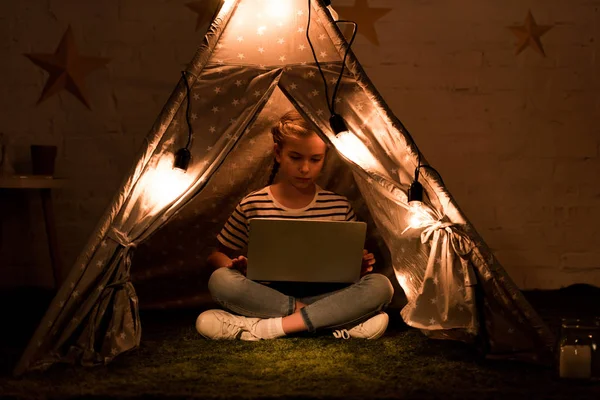 Concentrated kid using laptop in wigwam with luminous bulbs