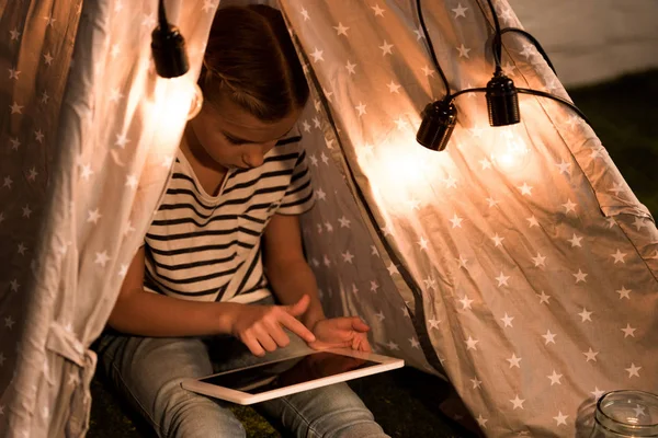 Kid in striped t-shirt sitting in wigwam and using digital tablet with blank screen