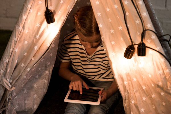 Child in striped t-shirt sitting in wigwam and using digital tablet with blank screen