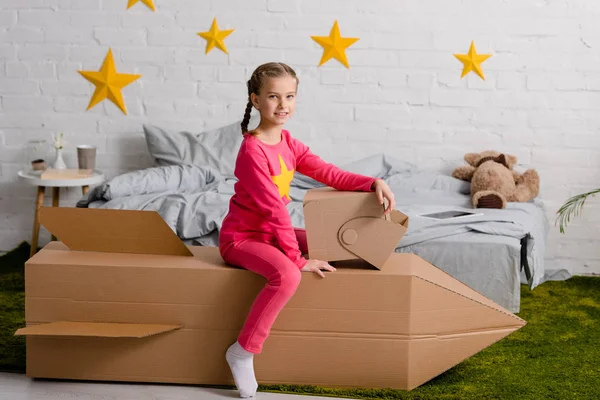 快乐的孩子与头盔坐在纸板火箭在卧室里 — 图库照片