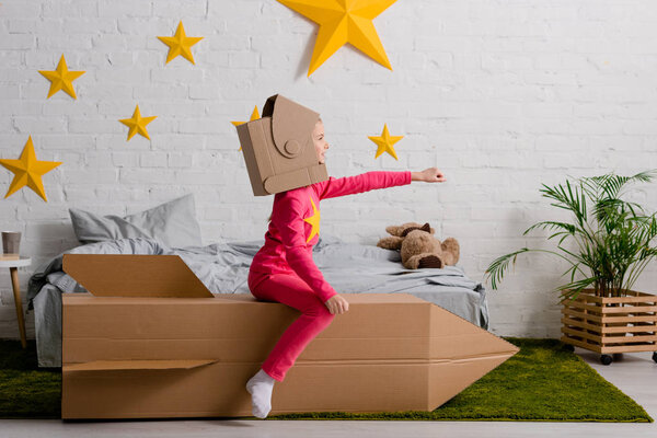 Funny kid in cardboard helmet sitting on rocket with hand up in bedroom