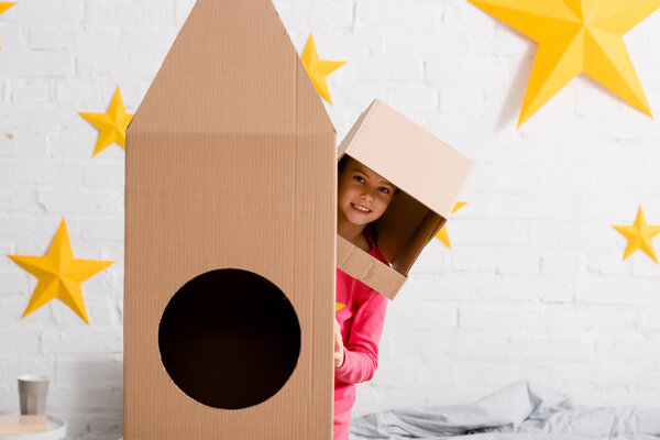 Funny child in cardboard helmet standing near rocket in bedroom