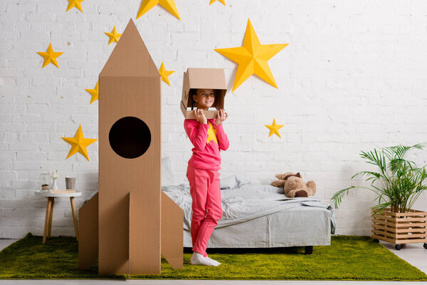 Вид в полный рост ребенка в шлеме, стоящего возле ракеты в спальне
