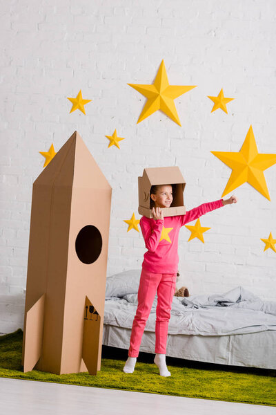 Full length view of kid in cardboard helmet standing near rocket in bedroom