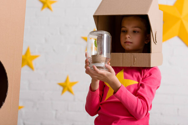 Curious child in cardboard helmet holding jar with plant