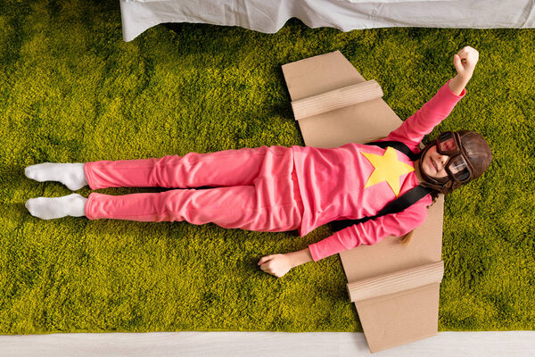 Top view of funny kid with cardboard wings lying on carpet with fist up
