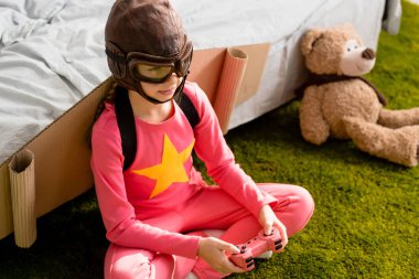 Child with cardboard wings sitting on carpet and playing video game with joystick clipart